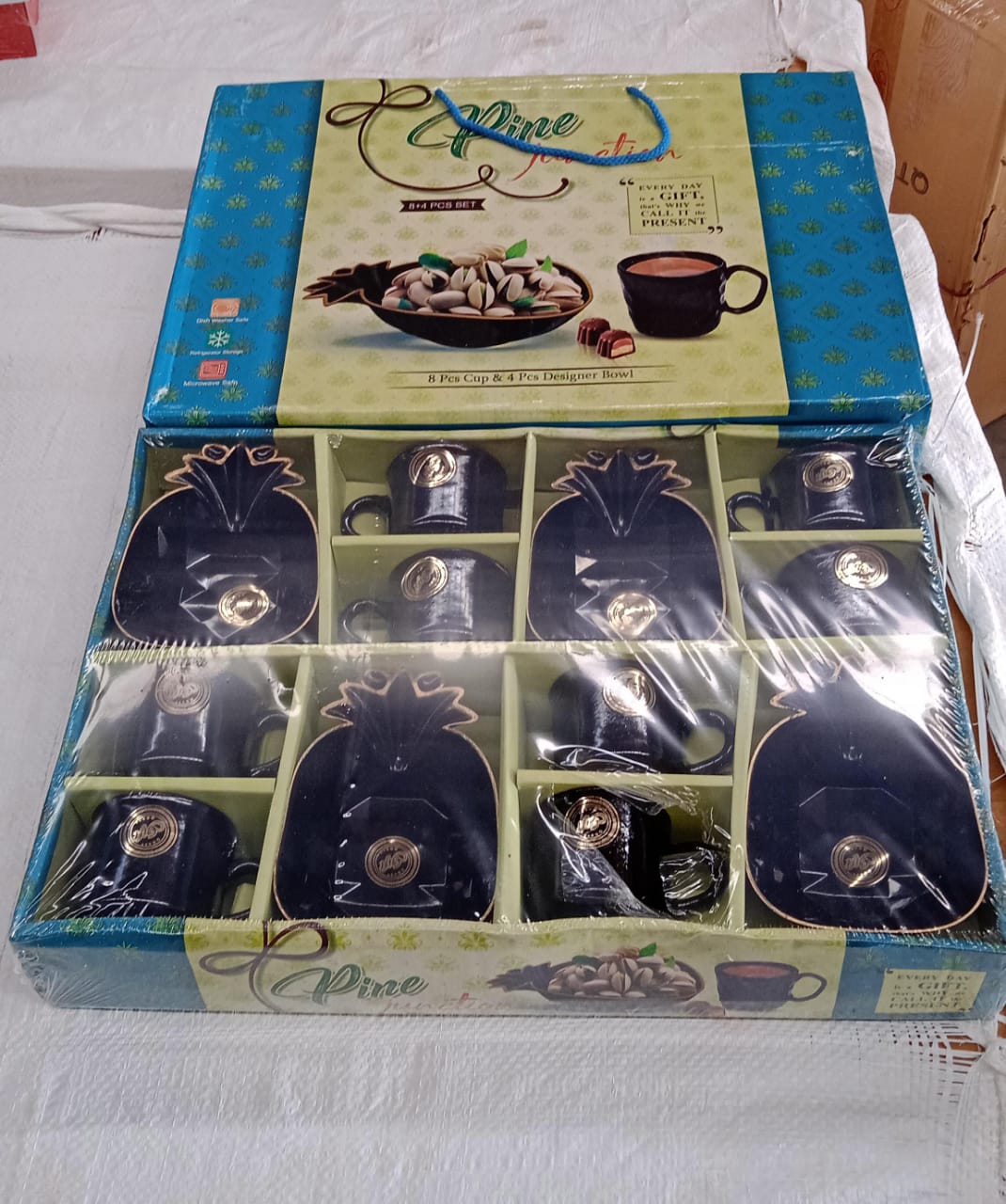 An open product box containing a black ceramic tea set with eight cups and four pineapple-shaped snack bowls, wrapped in