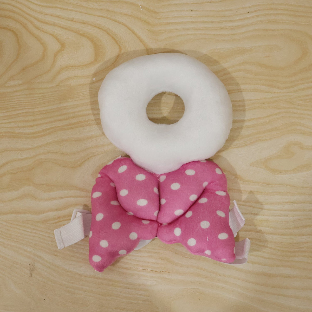 A pink and white polka dot head protector with angel wings lies flat on a light wooden surface.