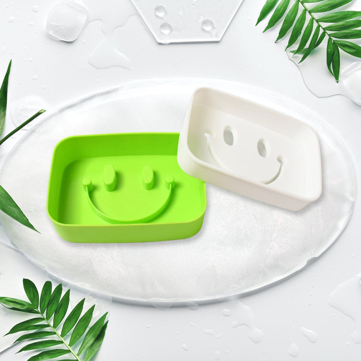 A green and a white soap dish, both with smiley face designs, on a white surface with green leaves.