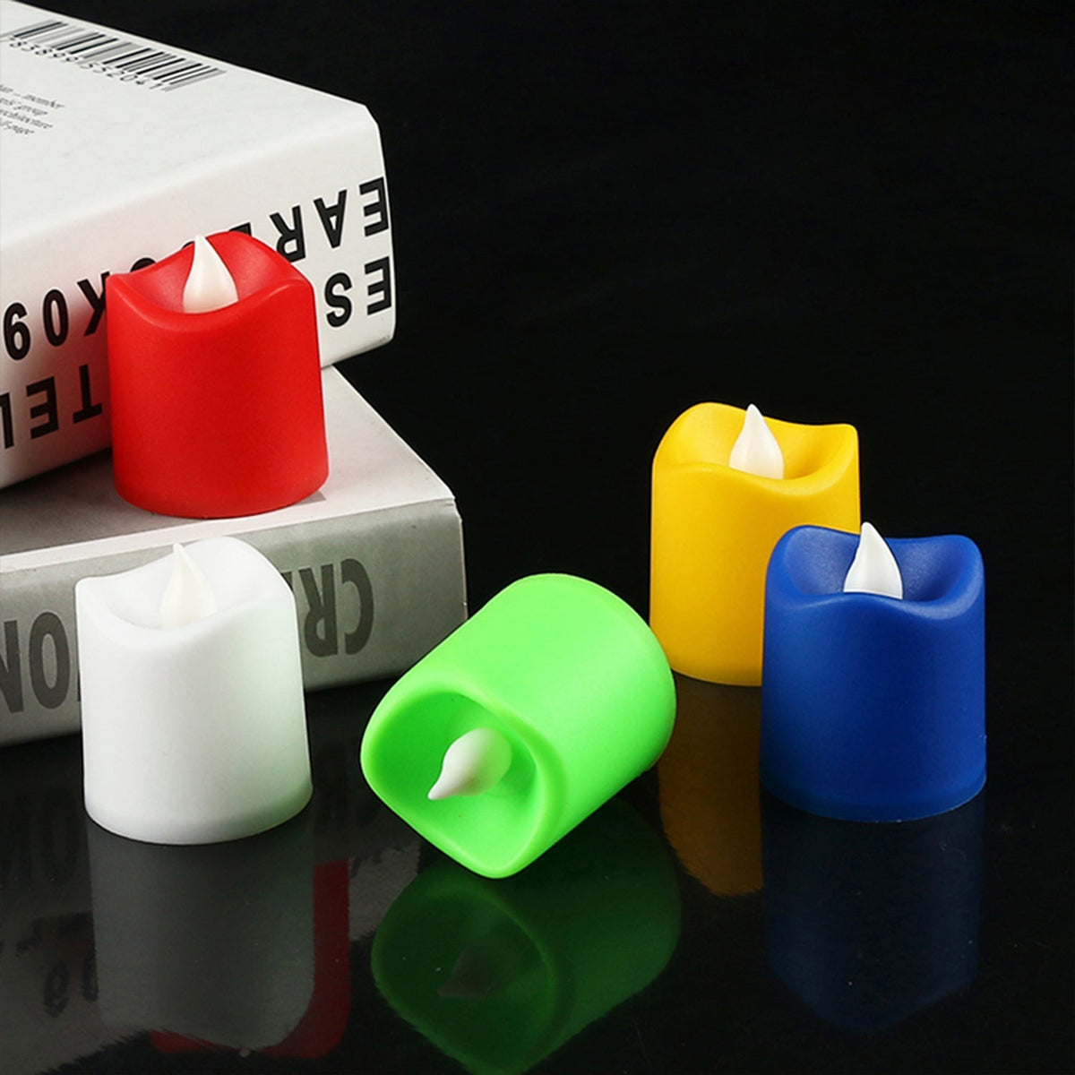 Five multicolor LED tealight candles, including white, red, green, yellow, and blue, placed on a dark surface next to a book.
