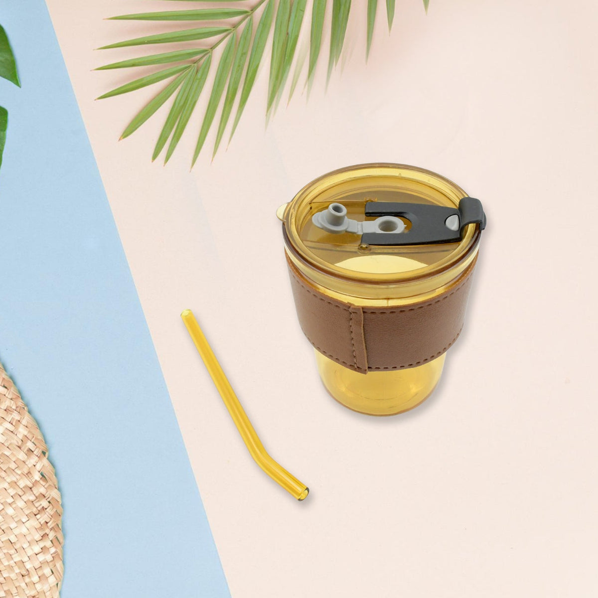 An amber glass cup with a brown leather sleeve and a separate straw are on a pink and blue background with palm leaves.