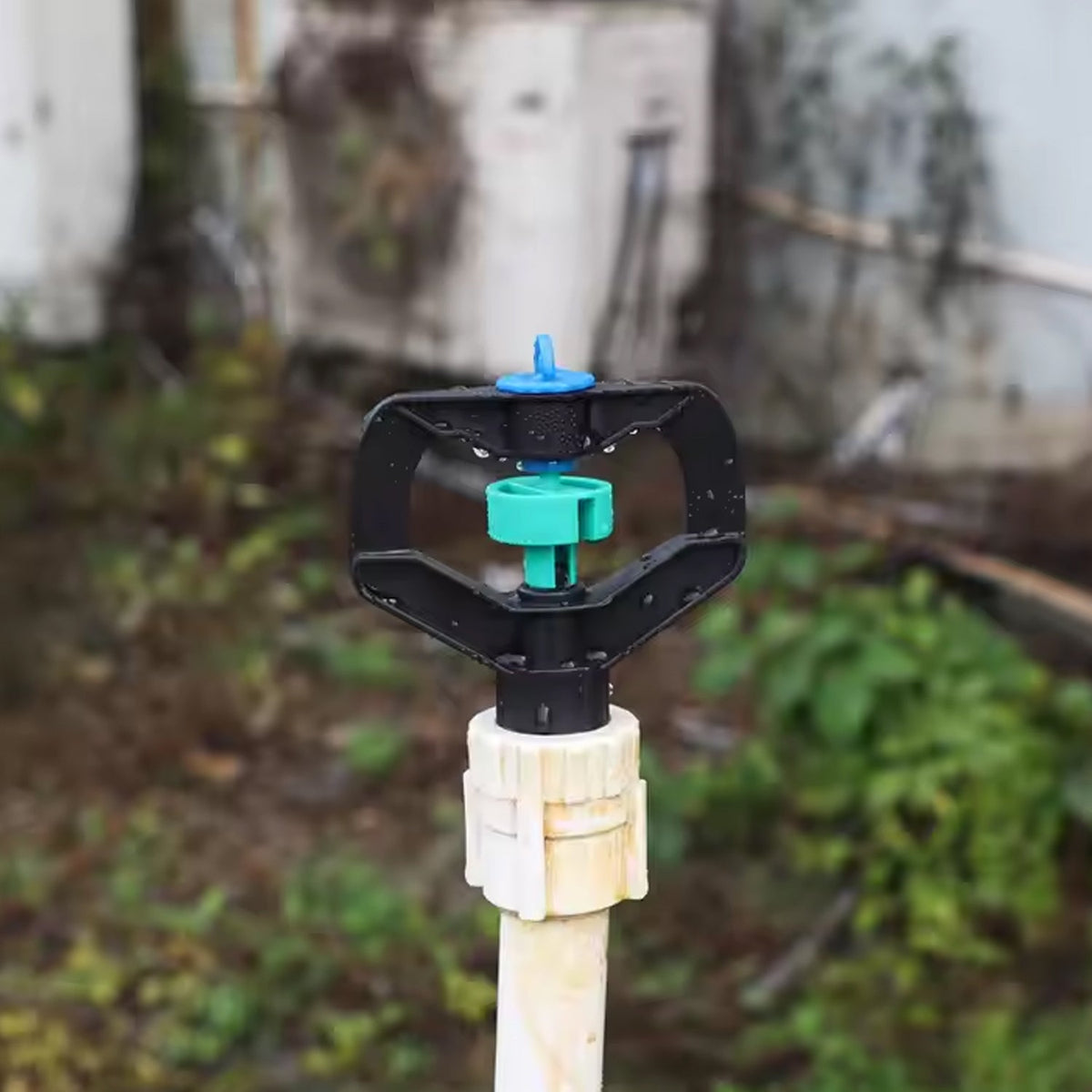 A black and green sprinkler nozzle attached to a white pipe, with a blurred garden background.