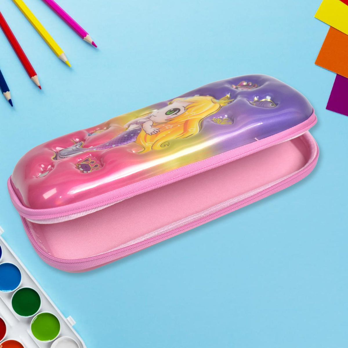 An open mermaid pencil case displaying its pink interior, with colorful pencils and watercolor paints in the background.