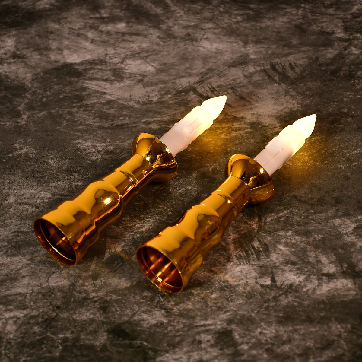 Two gold-based LED candles with lit flames lie diagonally on a textured dark gray surface.