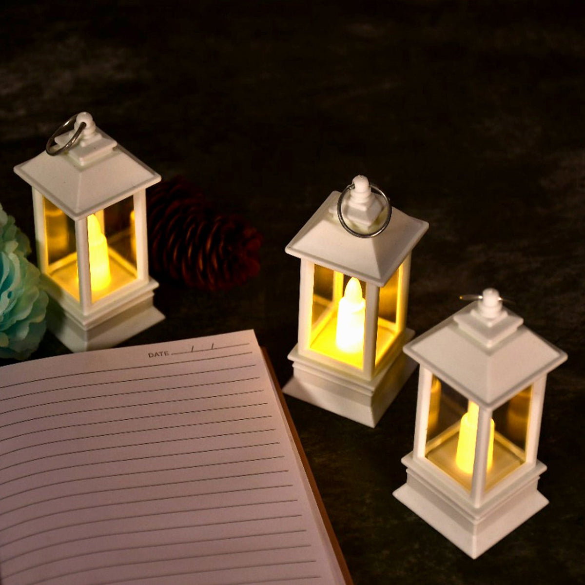 Three white lanterns with glowing LED candles and an open notebook on a dark surface, next to a teal flower and pinecone.