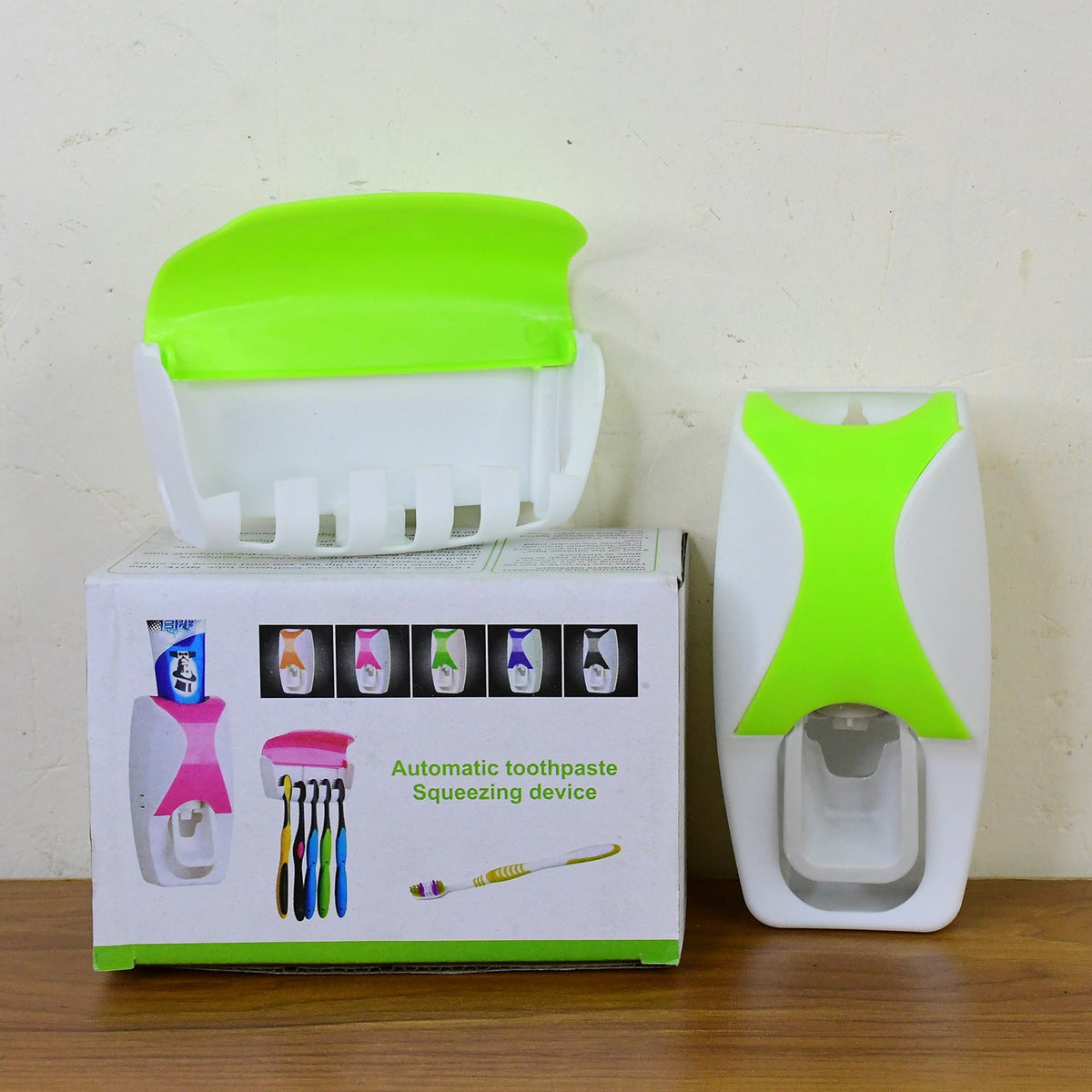The white and lime green automatic toothpaste dispenser, toothbrush holder, and product packaging box on a wooden surface.