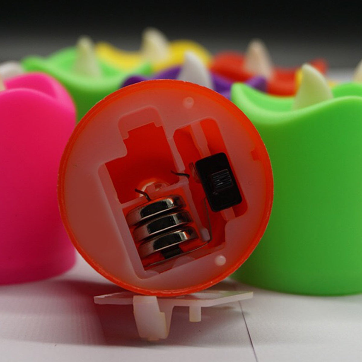 A close-up view of the underside of an orange LED tealight candle, revealing the battery compartment and switch.
