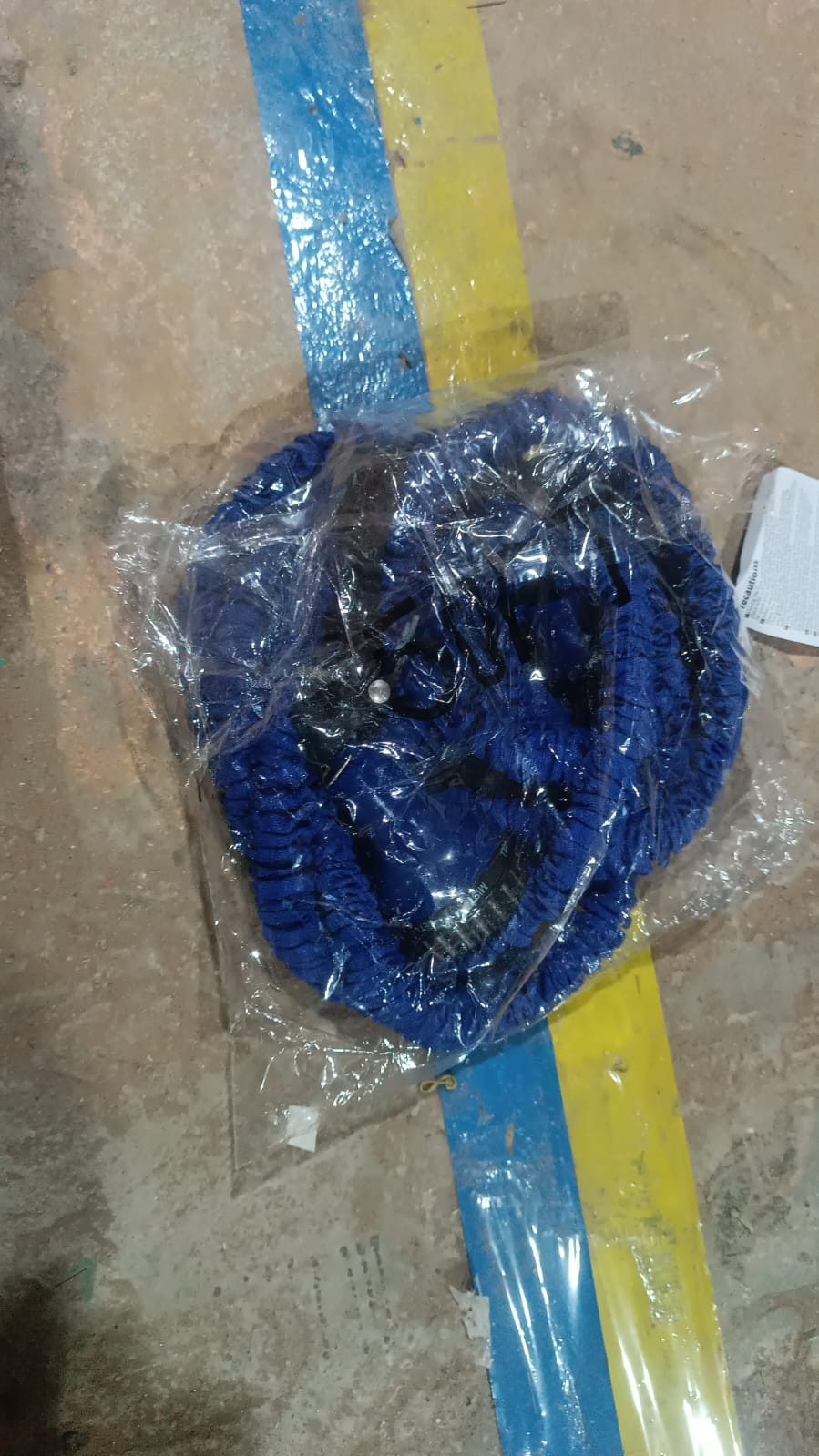 A blue expandable garden hose, coiled and sealed in clear plastic packaging, lying on a concrete floor with yellow and blue