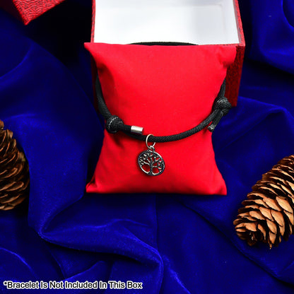 A black cord bracelet with a silver tree charm displayed on a red velvet pillow, surrounded by pinecones on a blue cloth.