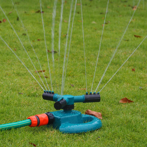 A teal and black garden sprinkler connected to a green hose waters a green lawn.