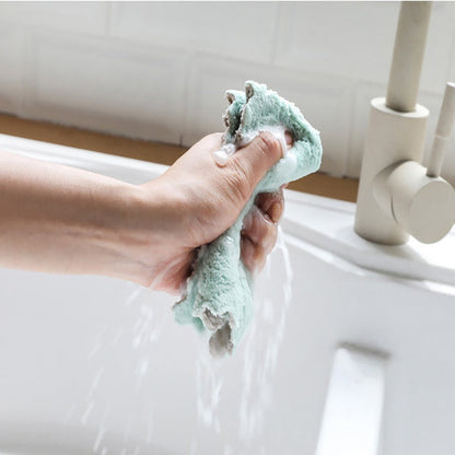 A hand firmly wrings out a light green microfiber towel under running water in a white kitchen sink.