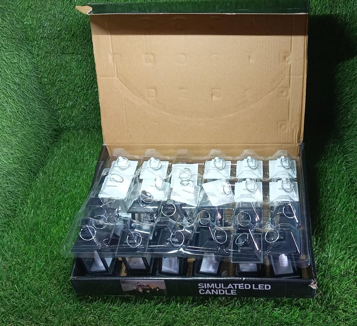 An open cardboard box containing twenty-four miniature black and white LED candle lanterns, sitting on a vibrant green grass