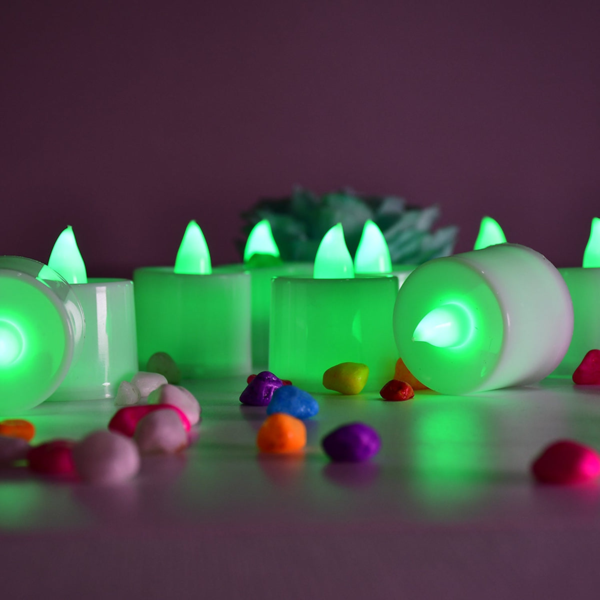 Several green flameless LED tealights are arranged on a surface with colorful pebbles, one tealight is on its side.