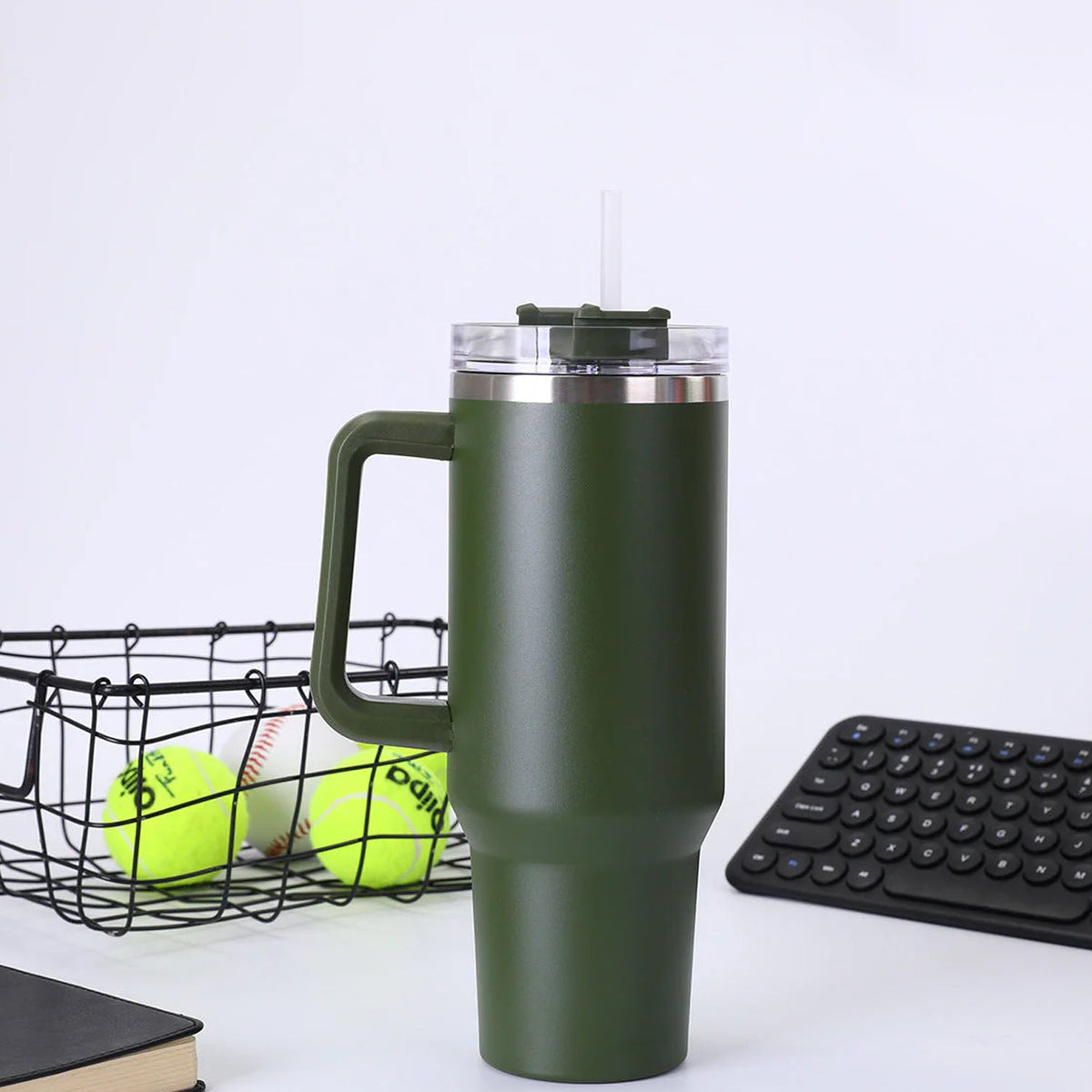 A dark green insulated travel mug with a clear lid and straw sits on a white desk next to a black wire basket with tennis