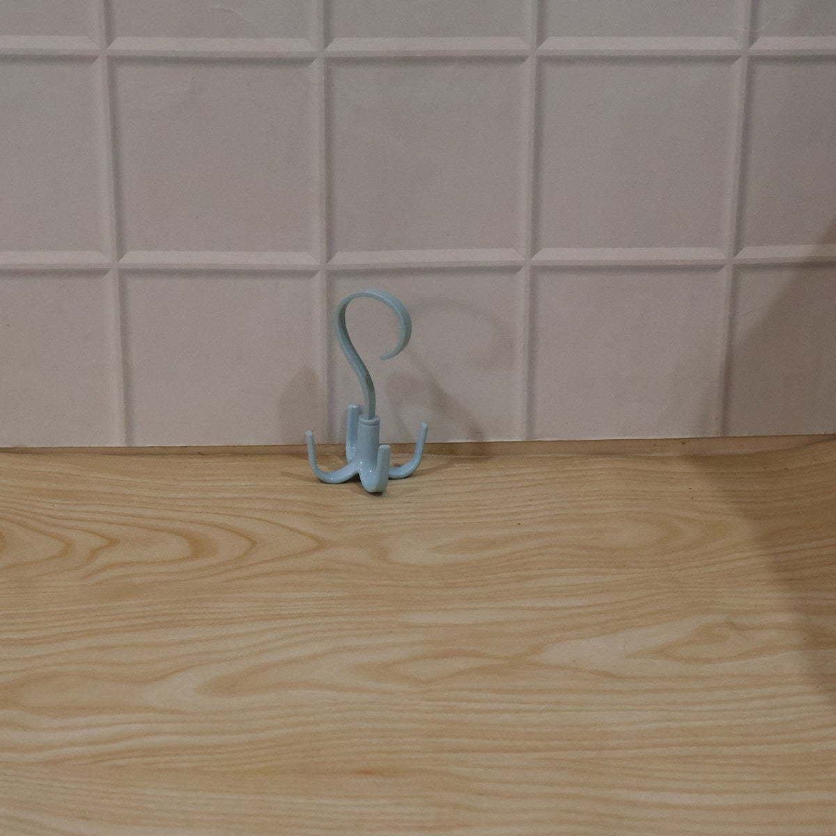 A light blue rotating multi-hook hanger sits on a light wooden surface against a white tiled wall.