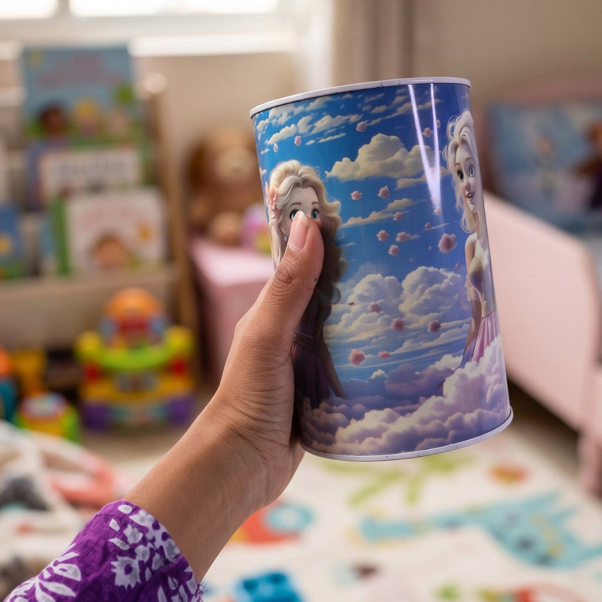 A hand holds the princess-themed money bank, showing two princesses among clouds, in a child's room.
