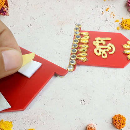 A hand peeling the backing off a red Shubh Labh wall hanging, revealing white adhesive pads, with scattered yellow and pink