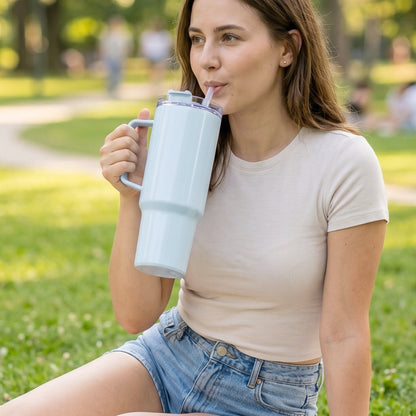 A woman sits on grass, sipping from a large light blue handled tumbler with a straw.