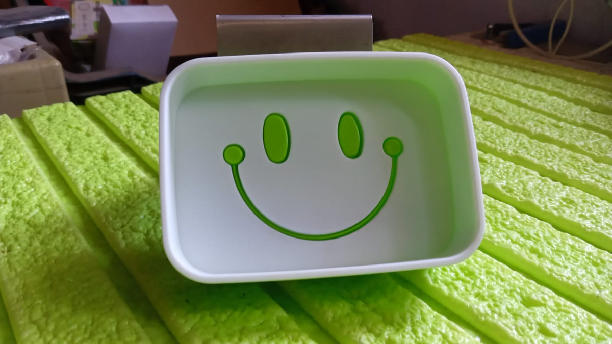 A green and white soap dish with a smiley face design, sitting on a textured green surface.